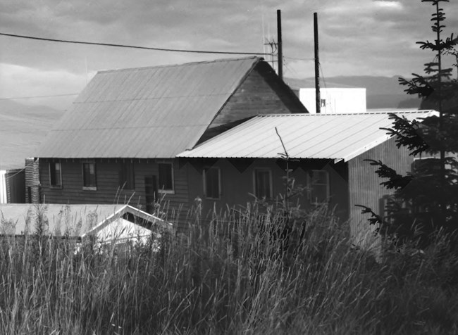 Tall grass grows in front of a long bunkhouse with a row of windows and metal roof