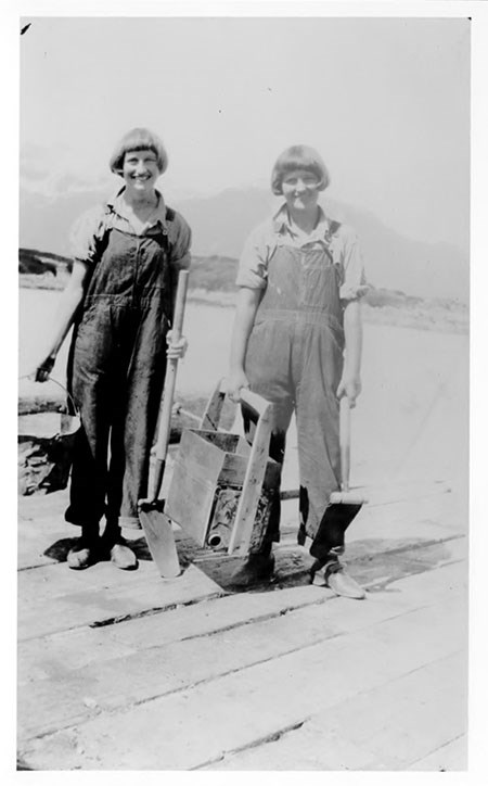 Two women with bobbed haircuts and overalls carry shovels and pails, black and white photo