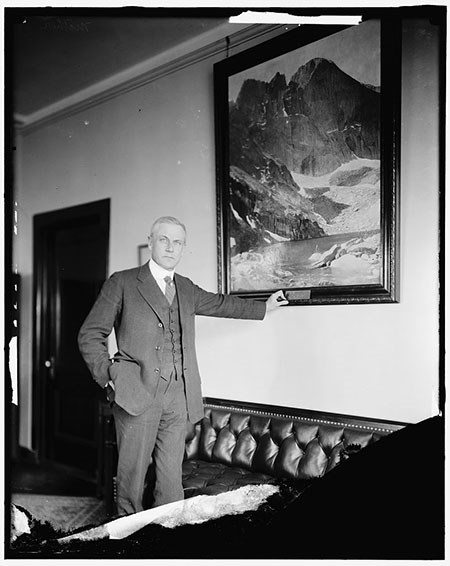 450-MatherinOffice-LOC A man in a suit and vest stands in an office next to a leather couch, his arm outstretched to the frame of a painting of a mountain.