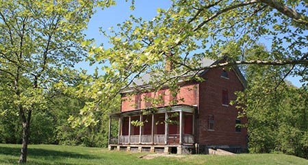 Leafy trees frame a view of a two-story red brick structure with a wooden porch across its full width and boarded windows.