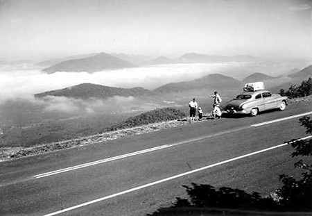 Four motorists stand behind a car parked alongside a highway, overlooking mountains interlaced with clouds.