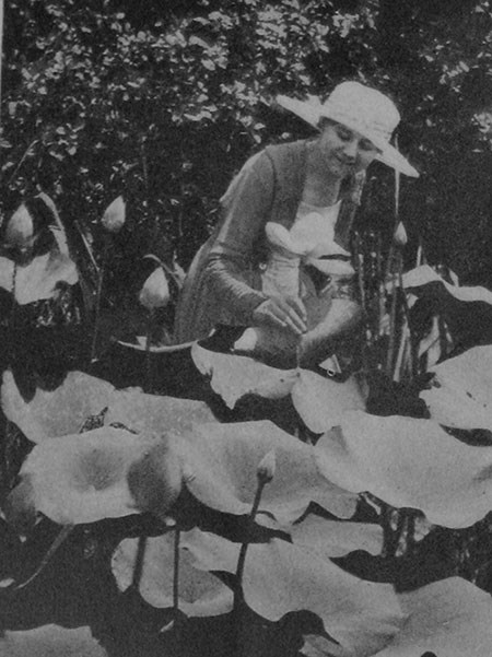 A woman in a wide-brimmed hat smiles as she touches a water lotus.