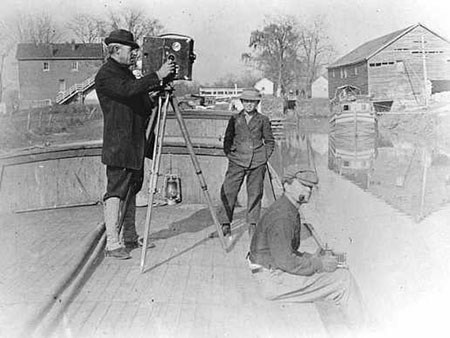 A man in a hat stands behind an early movie camera, balanced on a tripod on a canal boat.