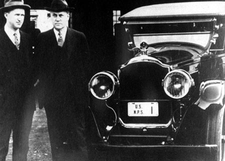 Two men in suits, ties, and brimmed hats stand near the front of a 1920s car.