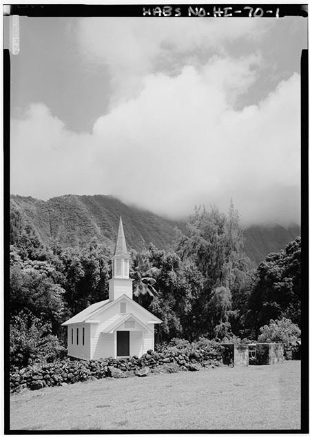 The sharp white spire of a small church stands out from dense foliage and low stone walls.