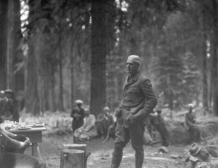 Stephen Mather, in uniform with high boots, buttoned jacket, and hands in pockets, stands amid onlookers in a forest.