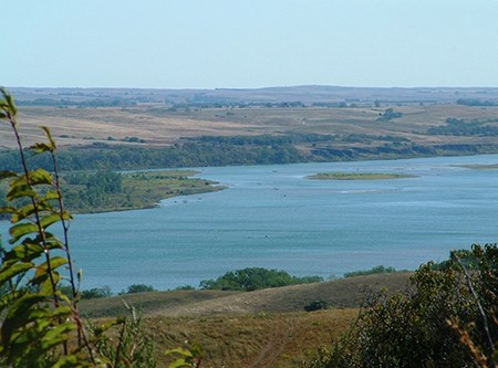 A wide blue river is framed by a rolling landscape of short, tan grass with clusters of trees