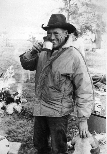450_frank-galey-standing A man in a cowboy hat and jacket smiles as he drinks from a tin cup near a fire.