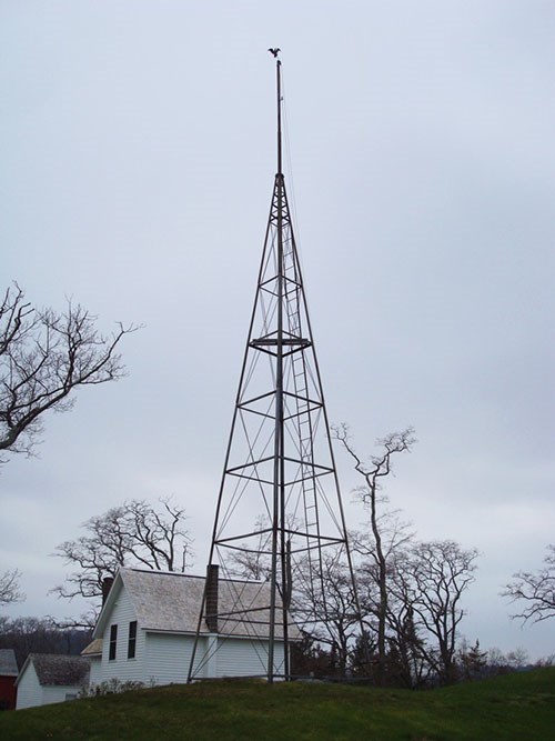 A metal signal tower rises to a point over a small building and bare trees.
