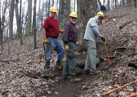 Three members of a trail crew with hardhats, work gloves, and tools, standing on the dirt trail surrounded by a forest of bare trees and leaf-covered ground.