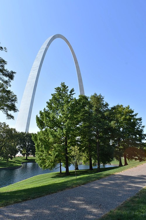 A path alongside a designed pond, encircled by trees and turf with the Gateway Arch rising in the background.