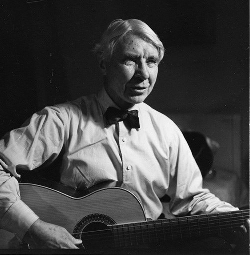 White-haired Carl Sandburg with a bow-tie and button-down shirt holds an acoustic guitar