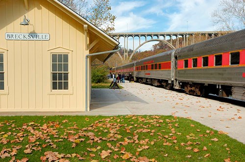 A train is stopped on a set of tracks beside a depot building that says "Brecksville." A tall bridge connects the hills in the background.