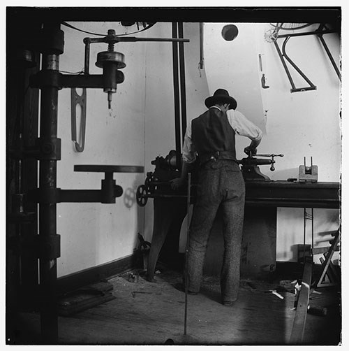 Wilbur Wright in hat and vest stands at a work bench in a workshop, back to camera, hand on a vise