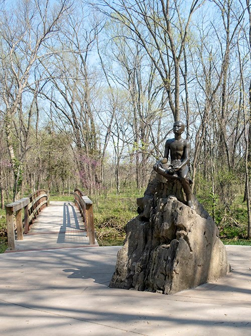 Sculpture of a young George Washington Carver seated on a rock, near a footbridge through spring woodlands