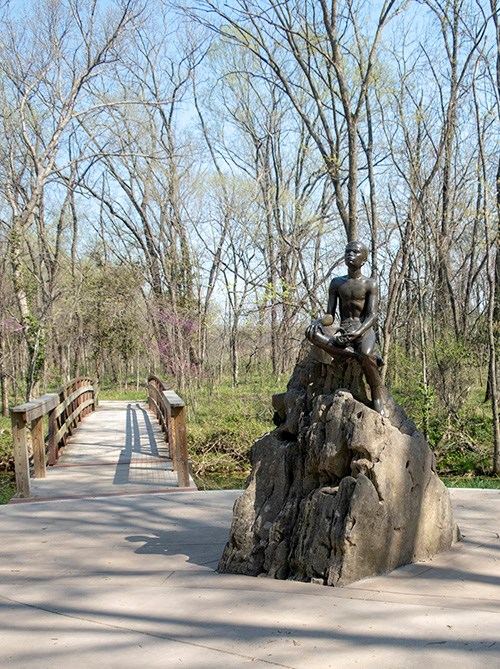 500-GWCA_Boy-Carver-Statue_040819_025-crop Sculpture of a young George Washington Carver seated on a rock, near a footbridge through spring woodlands