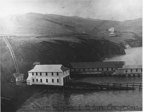 A black and white photograph of a two-story, white sided building with a boat ramp adjacent to a long, one-story building on a wharf, both stretching out from a steep-sided, hilly shoreline on the left into a bay on the right.