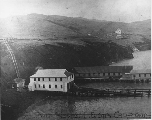 The Point Reyes Lifeboat Station's boathouse on Drakes Bay adjacent to the Paladini wharf with the chief's house in the background, ca. 1928. A black and white photograph of a two-story, white sided building with a boat ramp adjacent to a long, one-story building on a wharf, both stretching out from a steep-sided, hilly shoreline on the left into a bay on the right.