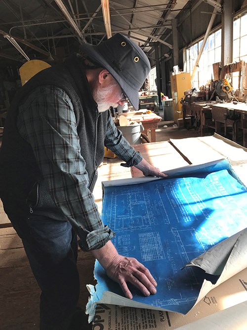 A man leans over a table in a warehouse over an unrolled blueprint