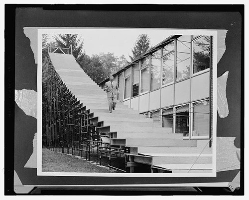A man in a suit descends a full-size model staircase next to a two story modern style building,