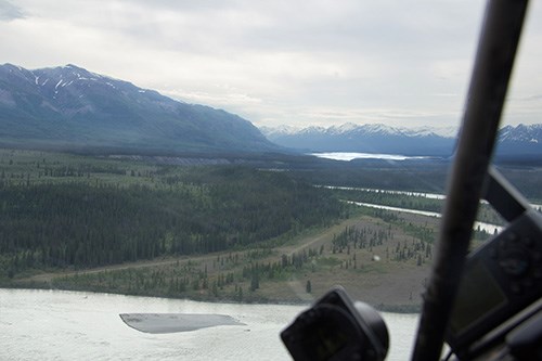 A rough airstrip is cleared between trees, glacier, and mountain, seen from a low plane.