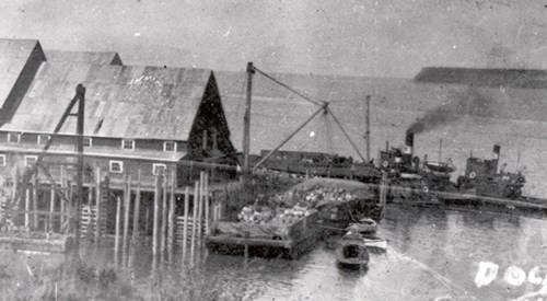 Grainy black and white image - various types of boat against a wooden dock, surrounding a warehouse with a steep roof