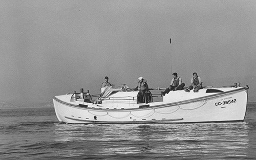 A black and white photo of four lifeboat station crew members aboard a 36-foot-long white lifeboat on gentle water.