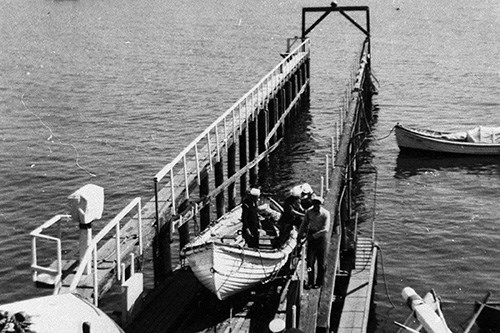 A black and white photograph of four lifeboat station crew members maneuvering a small, oar-powered lifeboat between the water and the boathouse along the boat launch's railway.
