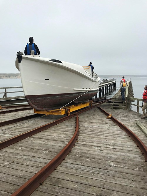 A color photograph of a 36-foot-long white- and red-painted lifeboat on a wheeled trolley being maneuvered along rail tracks down a boat launch into the water, as people in life vests watch and assist.