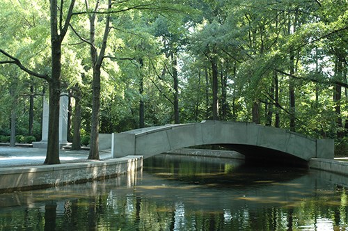 Water in a moat passes under a stone footbridge, near willow oaks