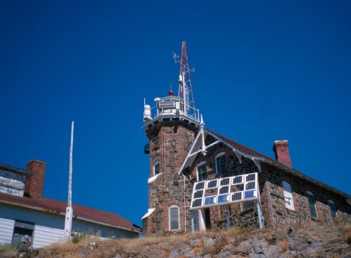 A short light house of red and brown brick stands on a rocky outcropping.
