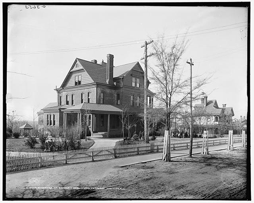 Young trees and shrubs and a fence surround a three-story Queen Anne style house