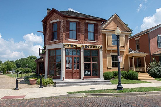 A two-story brick house with shutters stands on a corner of a brick street. A sign on the buildings says "The Wright Cycle Co."