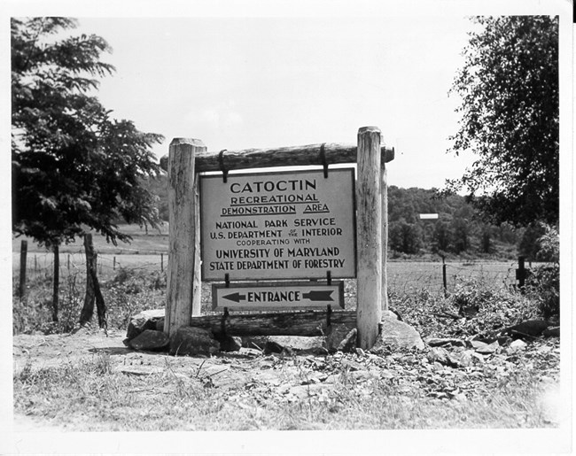 A sign on a wooden frame says "Catoctin Recreational Demonstration Area" with an arrow to the entrance, in a rural landscape.