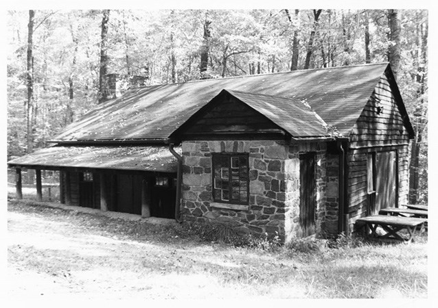 A rustic cabin made of wood and stone panels with wood log pillars. The cabin sits in front of a stand of deciduous trees.