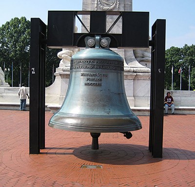 The bronze bell with sea foam colored patina hovers over a circular red brick pattern with white marble gleaming in the background.