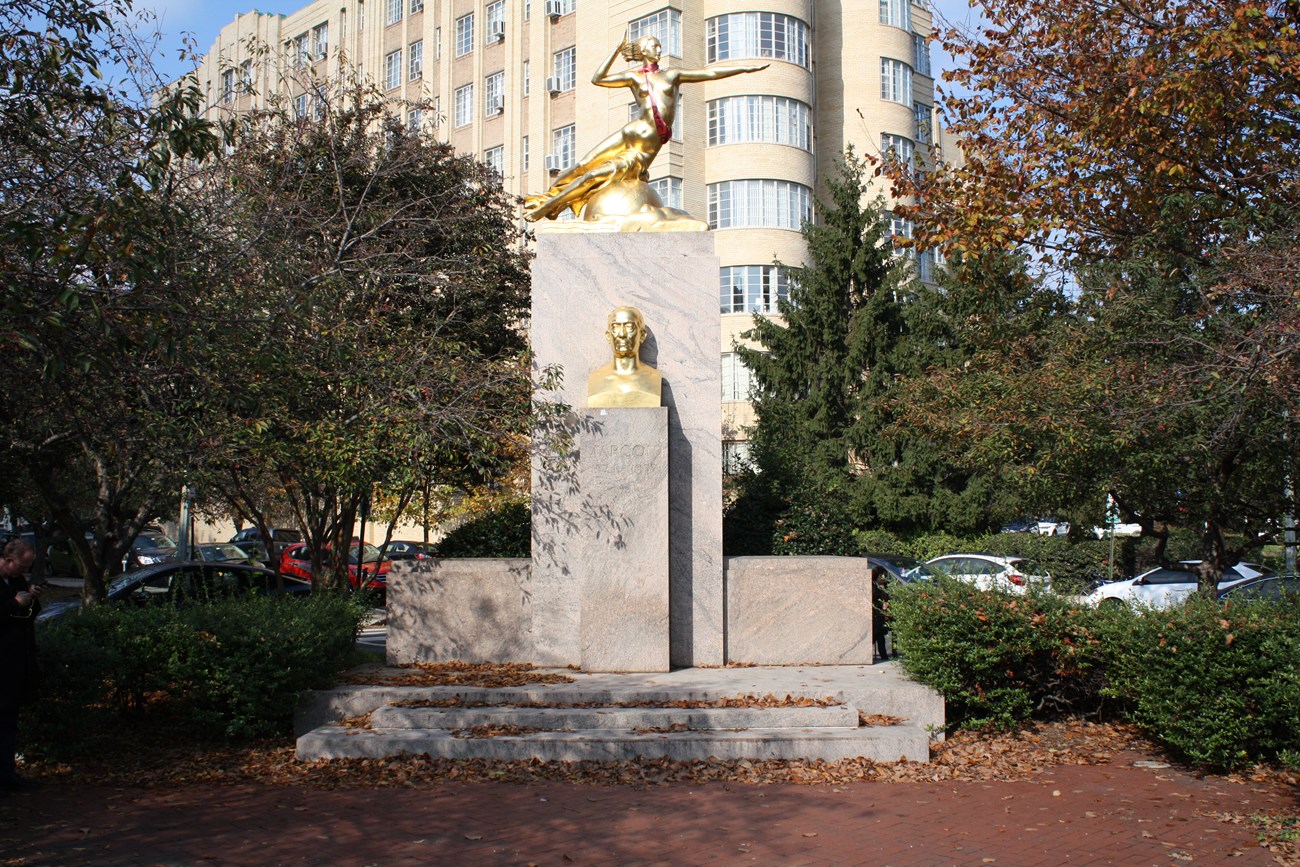 The pink granite and gilded bronze shine in the sun, framed by trees and shrubs on either side.