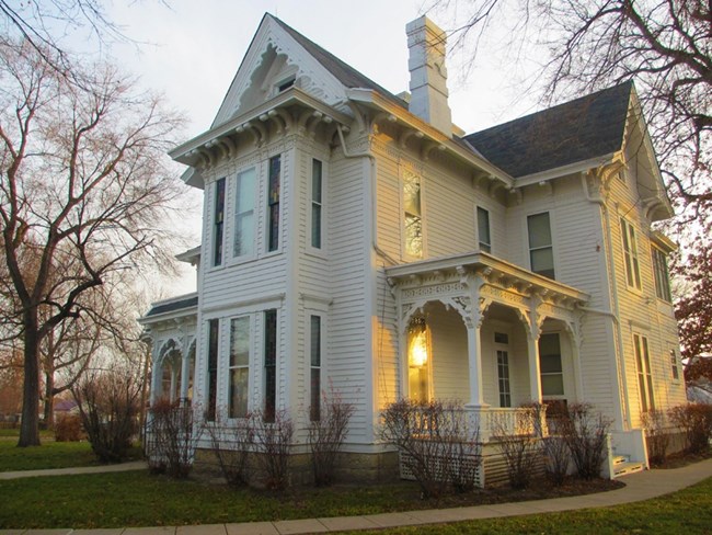 Leafless shrubs grow at the foundation of a Victorian house, framed by tall leafeless trees