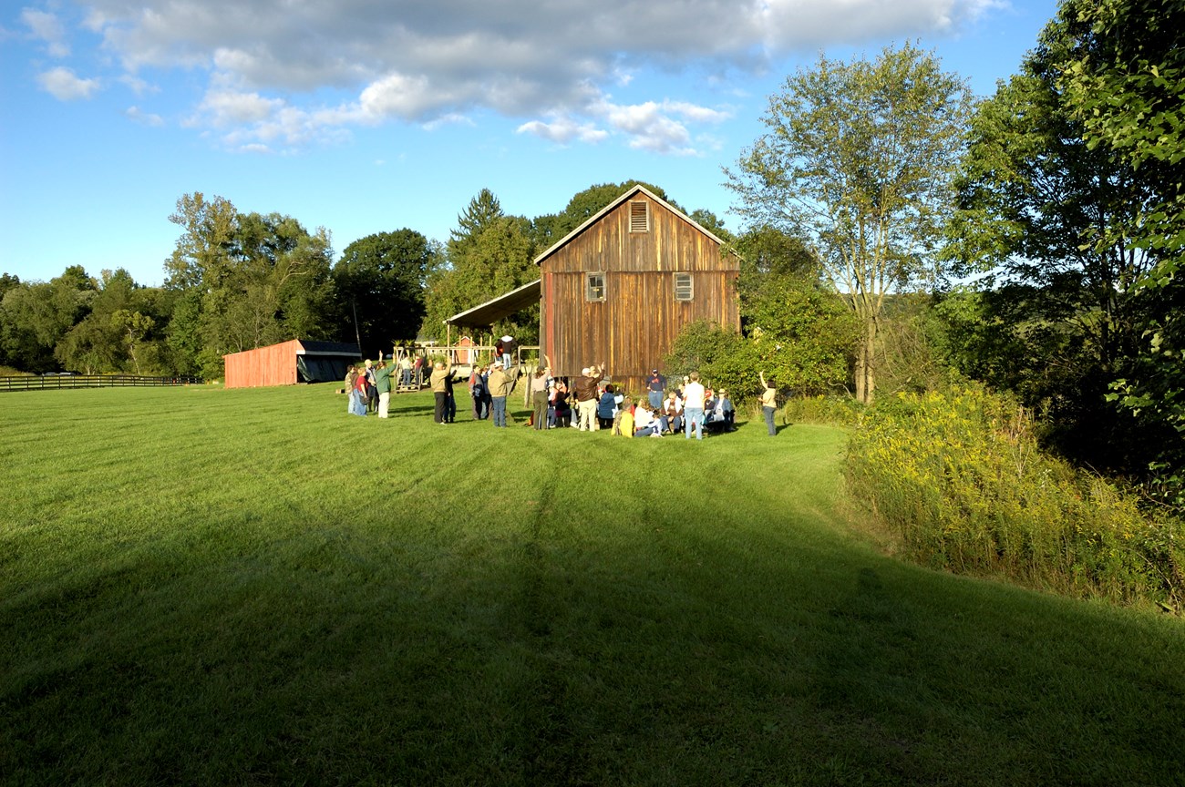A group of people stands in mowed grassy area beside a large barn