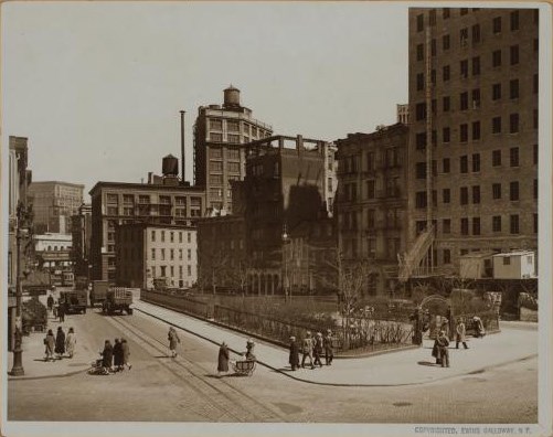 People walk on a sidewalk and in the street beside a small city park surrounded by buildings, 1928.