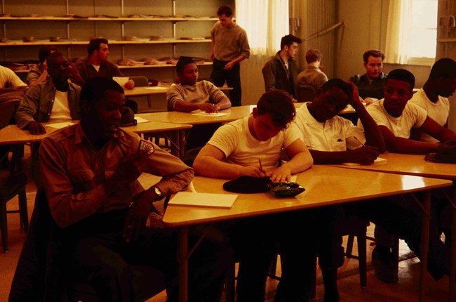 Young men sit in rows of tables with pencils and tablets