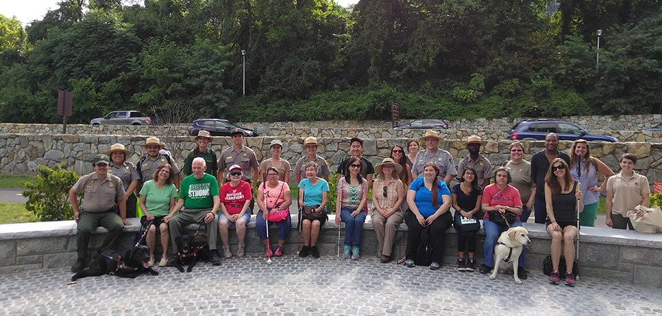 Two rows of participants - including park rangers, some visually impaired individuals, and several dogs - line up along a stone wall for a photo.