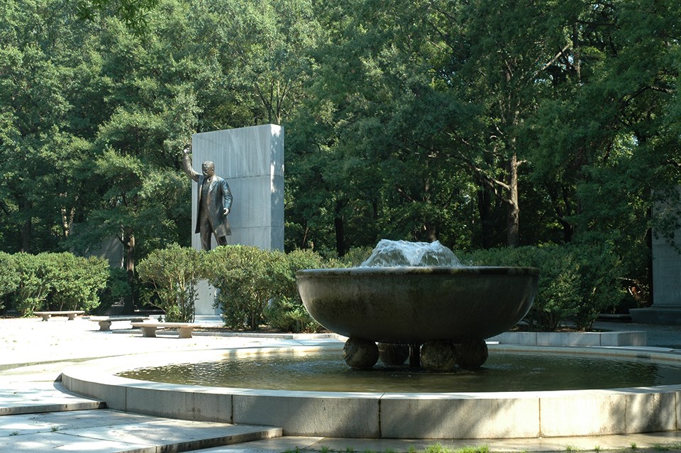 Water sprays out of a bowl fountain in a plaza, with a statue of Roosevelt behind