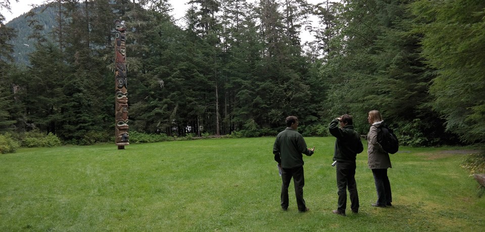 Three people stand in an area of grass surrounded by trees, a totem pole, and mountains.