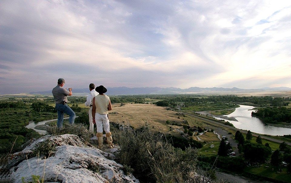 Three individuals face away, observing a river valley from an overlook