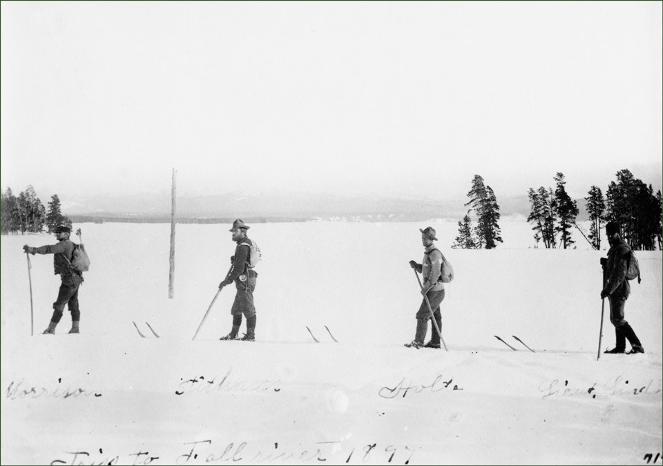 Four men on skis cross an open area of snow in 1897.