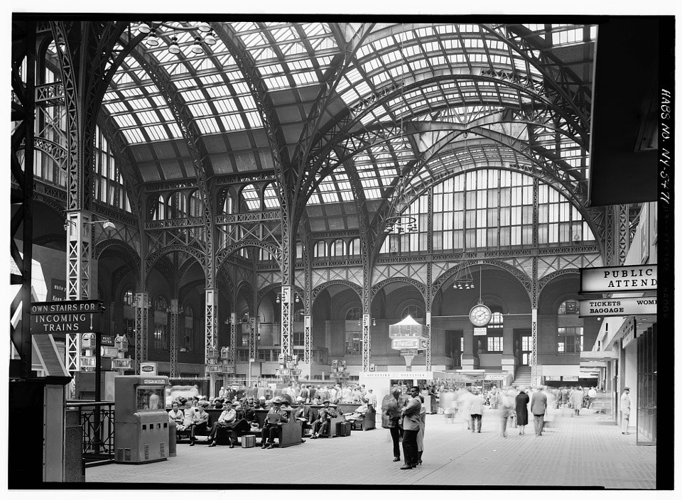 Passengers gather in the expansive concourse of a train station under an arching glass and metal ceiling.