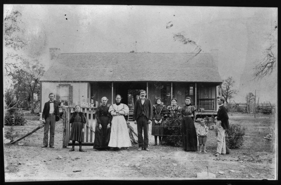 A family of ten poses along a fence in a dirt yard in front of the Johnson family home.