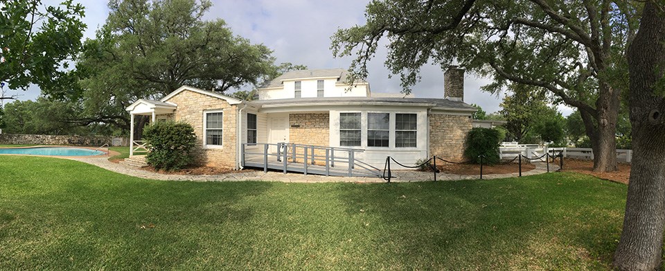 The inground pool is to the left of the ranch house, surrounded by a neat lawn and tall oaks.