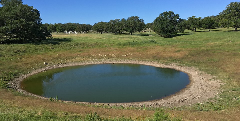 A still, round water pool is surrounded by a gently-rolling agricultural landscape.